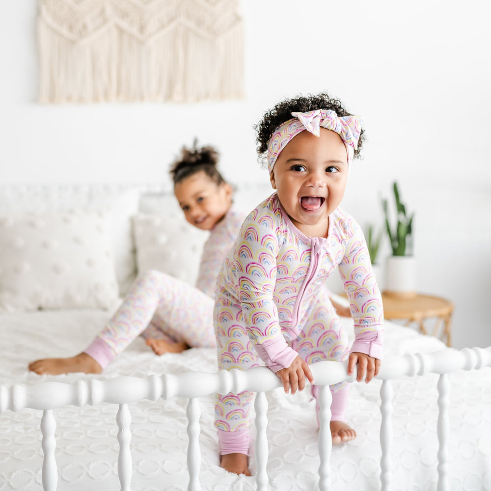 Image of infant girl wearing rainbow printed zip up romper with matching bow headband. This print features multicolored rainbows that sit upon a white background with light pink accent trim.