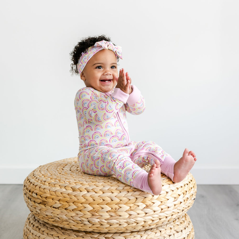Image of infant girl wearing rainbow printed zip up romper with matching bow headband. This print features multicolored rainbows that sit upon a white background with light pink accent trim.